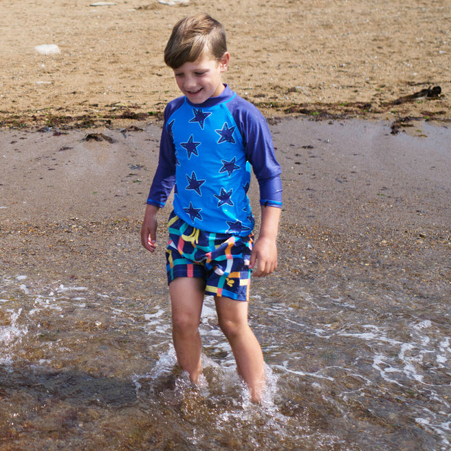 A young boy wearing a UV Protective Rash Vest Blue Starfish is standing in the sea with the waves crashing around his legs.